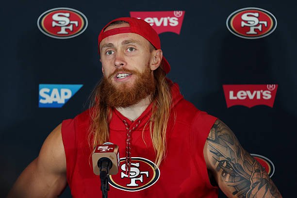 George Kittle speaks into a microphone wearing a red 49ers sleeveless hoodie and a backward red cap.