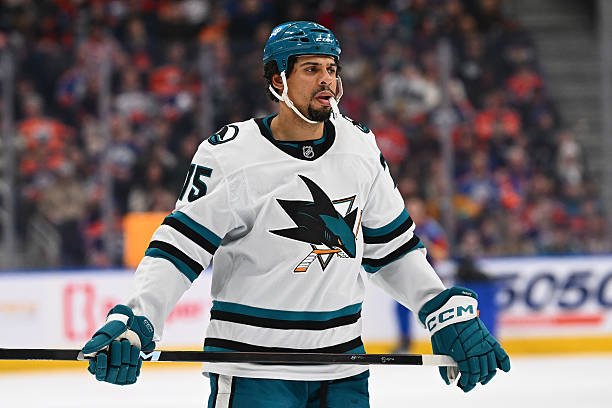 Ryan Reaves in a white San Jose Sharks jersey and teal helmet, holding his stick on the ice.