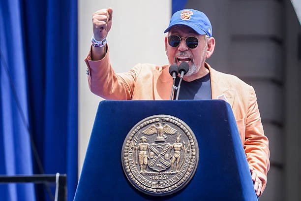 James Dolan wearing a Knicks cap and raising his fist while speaking at a podium.