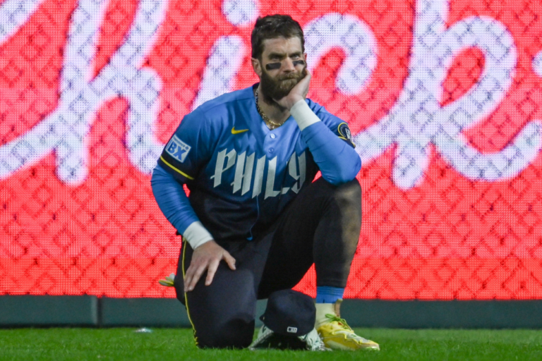 Bryce Harper kneels on the field in a blue Phillies training top.