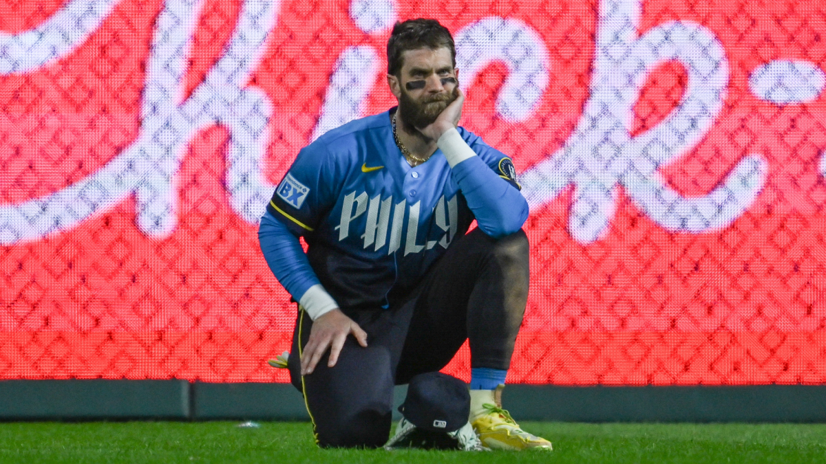 Bryce Harper kneels on the field in a blue Phillies training top.