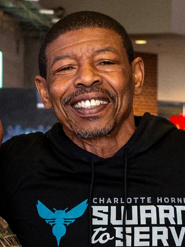 Muggsy Bogues smiles while wearing a Charlotte Hornets sweatshirt.