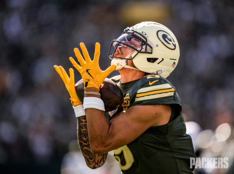 Packers receiver Christian Watson holds a football while looking upward at Lambeau Field.