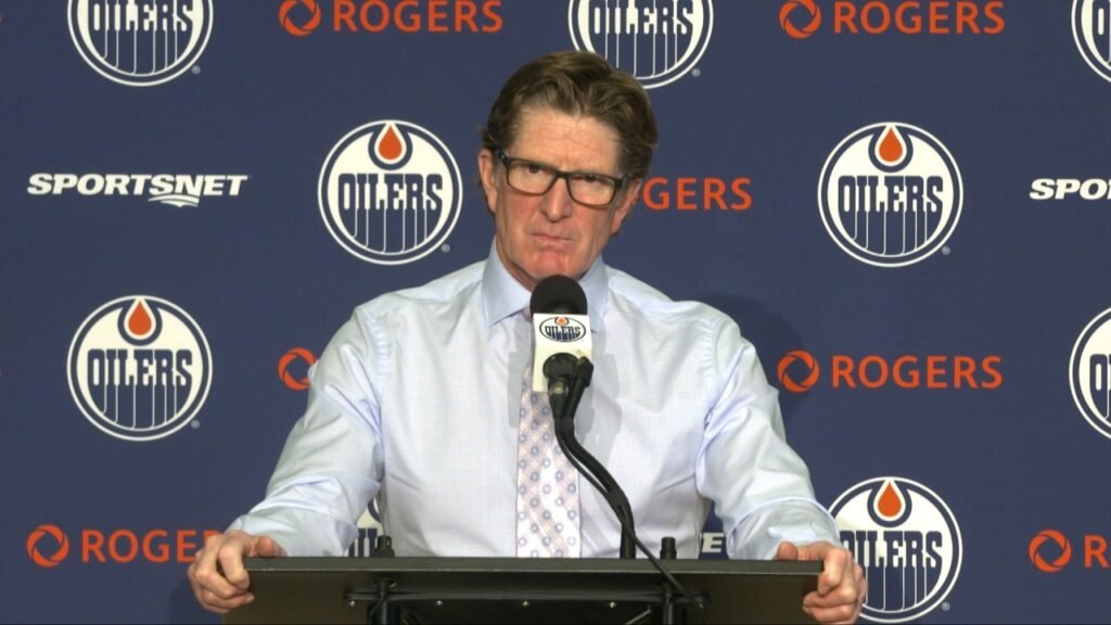 Mike Babcock speaks at a microphone in front of an Oilers backdrop.