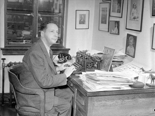 Foster Hewitt sitting at an office desk beside a typewriter and telephone.