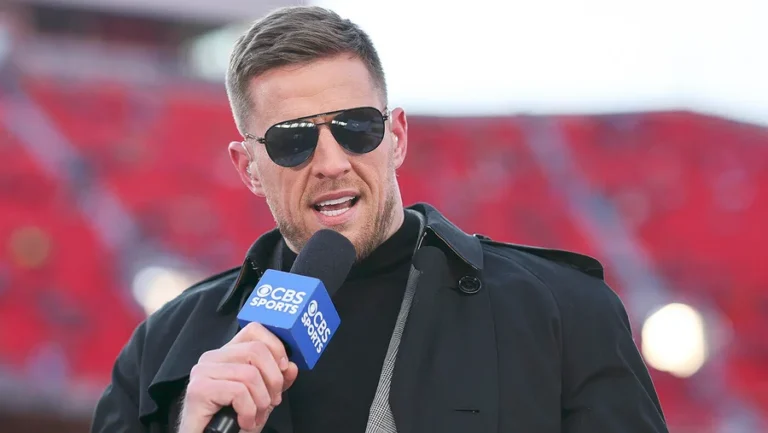 J.J. Watt wearing sunglasses and a black jacket while holding a microphone at a stadium.