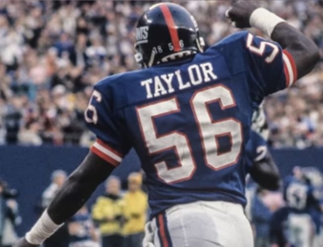 Lawrence Taylor in a blue New York Giants number 56 jersey with his right arm raised.