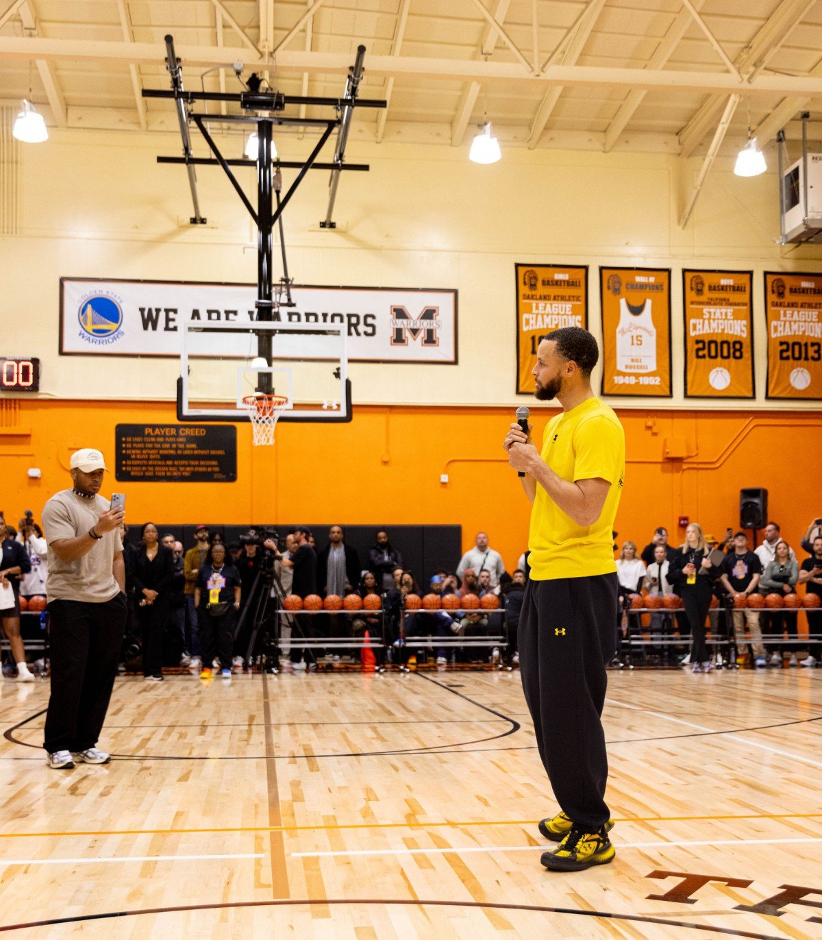 Stephen Curry in a yellow shirt holding a microphone on a basketball court.