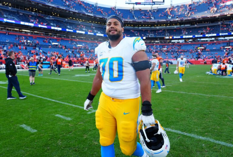 Rashawn Slater in a white Chargers jersey and yellow pants holding his helmet on the field.