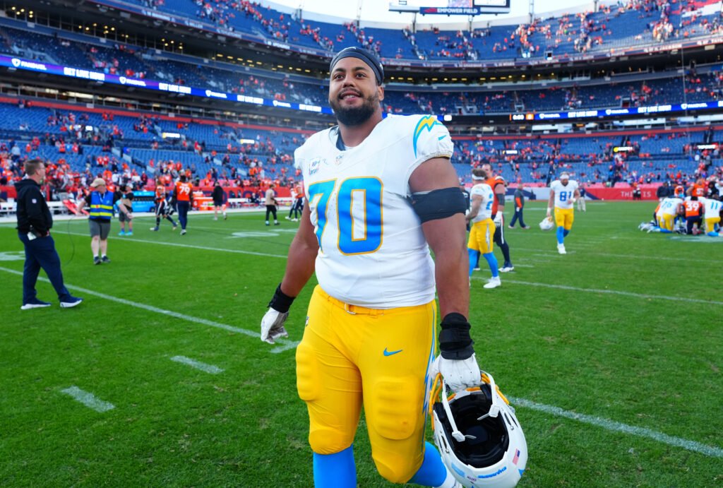 Rashawn Slater in a white Chargers jersey and yellow pants holding his helmet on the field.