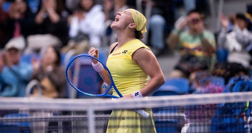 Diana Shnaider celebrates beside the net in a yellow tennis outfit.
