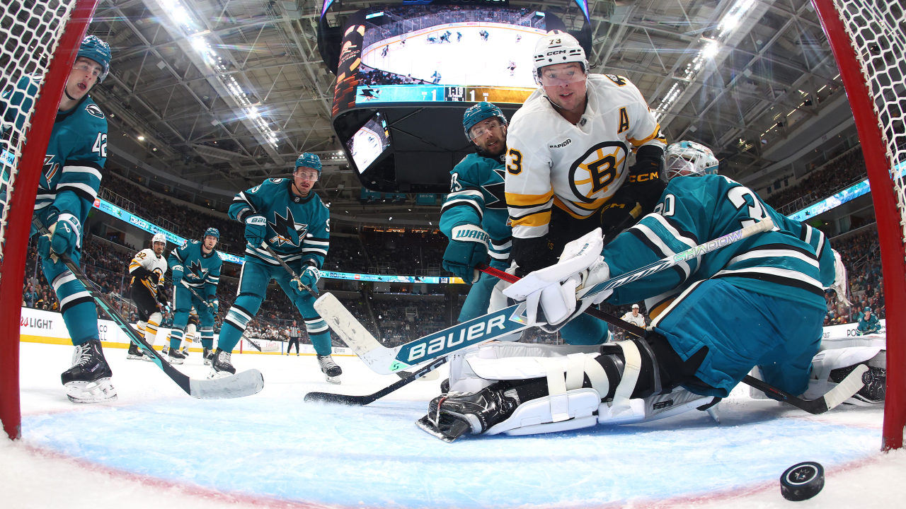 A Boston Bruins player battles in front of the San Jose Sharks goalie, viewed from inside the goal.