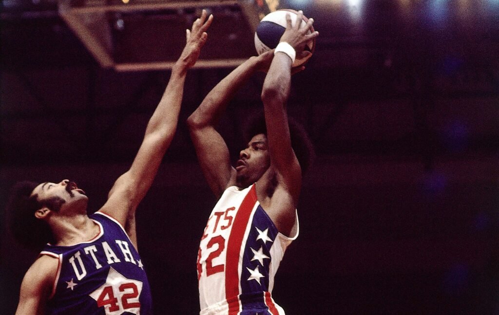 Julius Erving in a red Nets No. 32 jersey shoots over a Utah defender with the ABA’s red, white and blue ball.