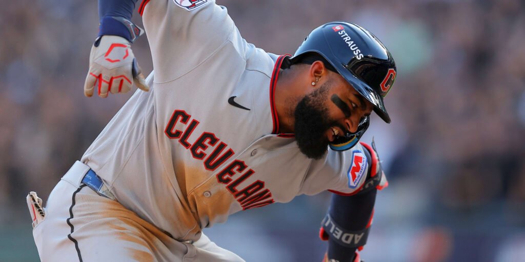 Jo Adell in a gray Cleveland Guardians uniform and batting helmet, leaning forward with his arm raised.