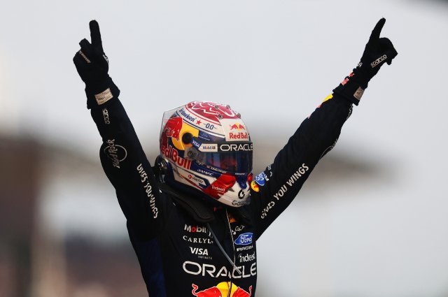 Max Verstappen raises both arms while wearing his Red Bull race suit and helmet.