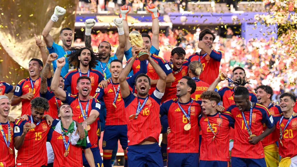 Spain players celebrate with the World Cup trophy on a podium amid gold confetti.