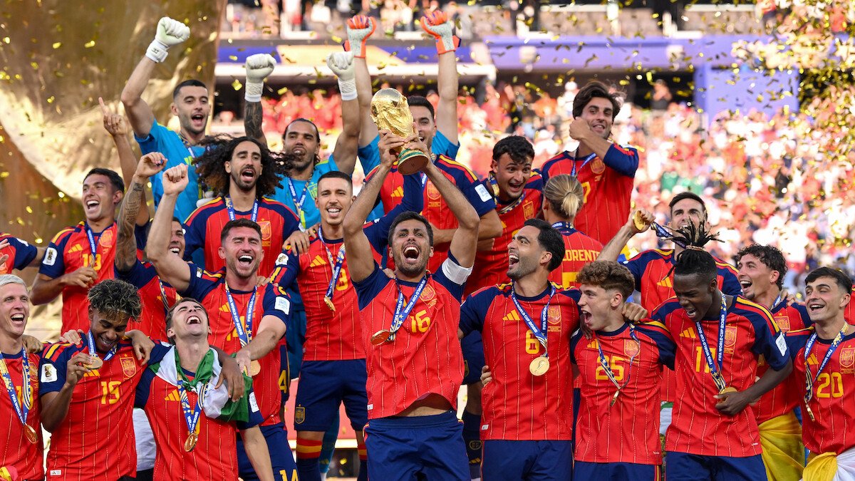 Spain players celebrate with the World Cup trophy on a podium amid gold confetti.