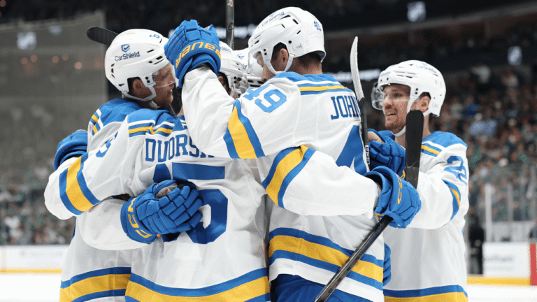 St. Louis Blues players celebrate together in white road uniforms beside the boards.