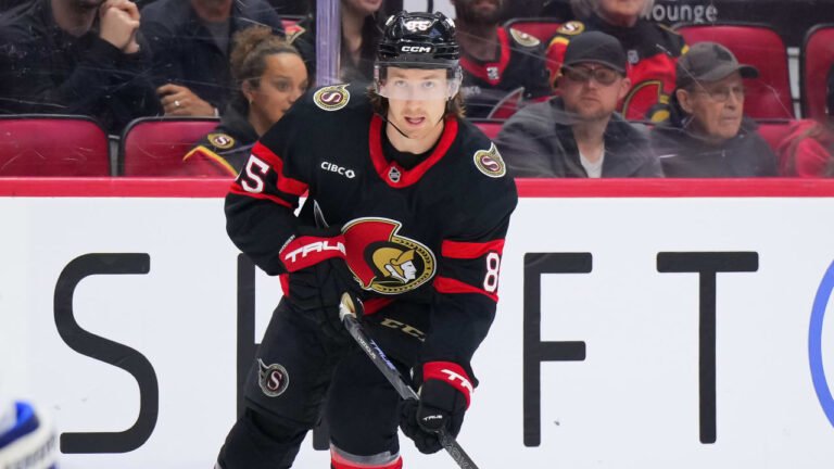 Jake Sanderson handles his stick in Ottawa’s black No. 85 uniform in front of the boards.