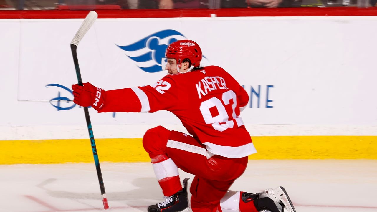 Marco Kasper celebrating on one knee in a red Detroit jersey, holding his stick.