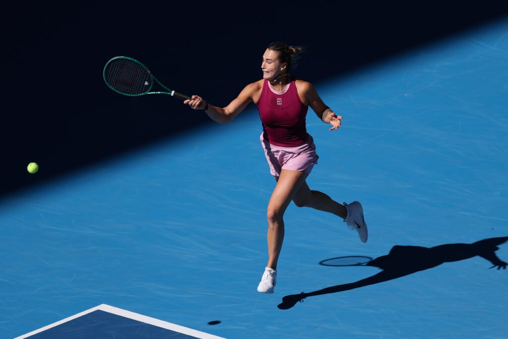 Aryna Sabalenka reaches forward to play a forehand on a blue hard court.